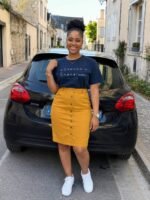 tshirtchretienfemmenecrainspasbleumarine Woman posing in a navy graphic T shirt mustard skirt and white sneakers on a cobblestone street beside a black hatchback smiling at the camera