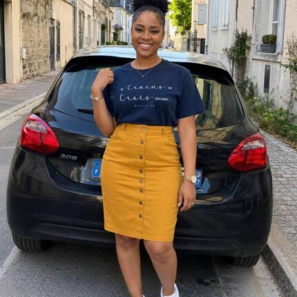 Woman posing in a navy graphic T-shirt, mustard skirt, and white sneakers on a cobblestone street beside a black hatchback, smiling at the camera.