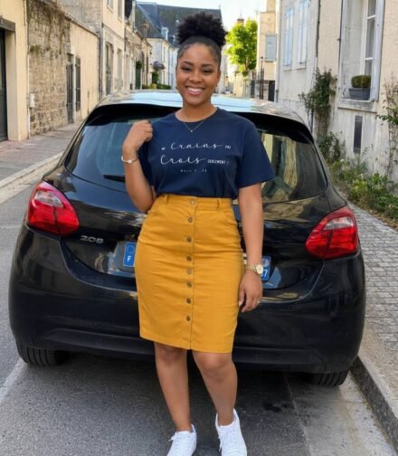 Woman posing in a navy graphic T-shirt, mustard skirt, and white sneakers on a cobblestone street beside a black hatchback, smiling at the camera.