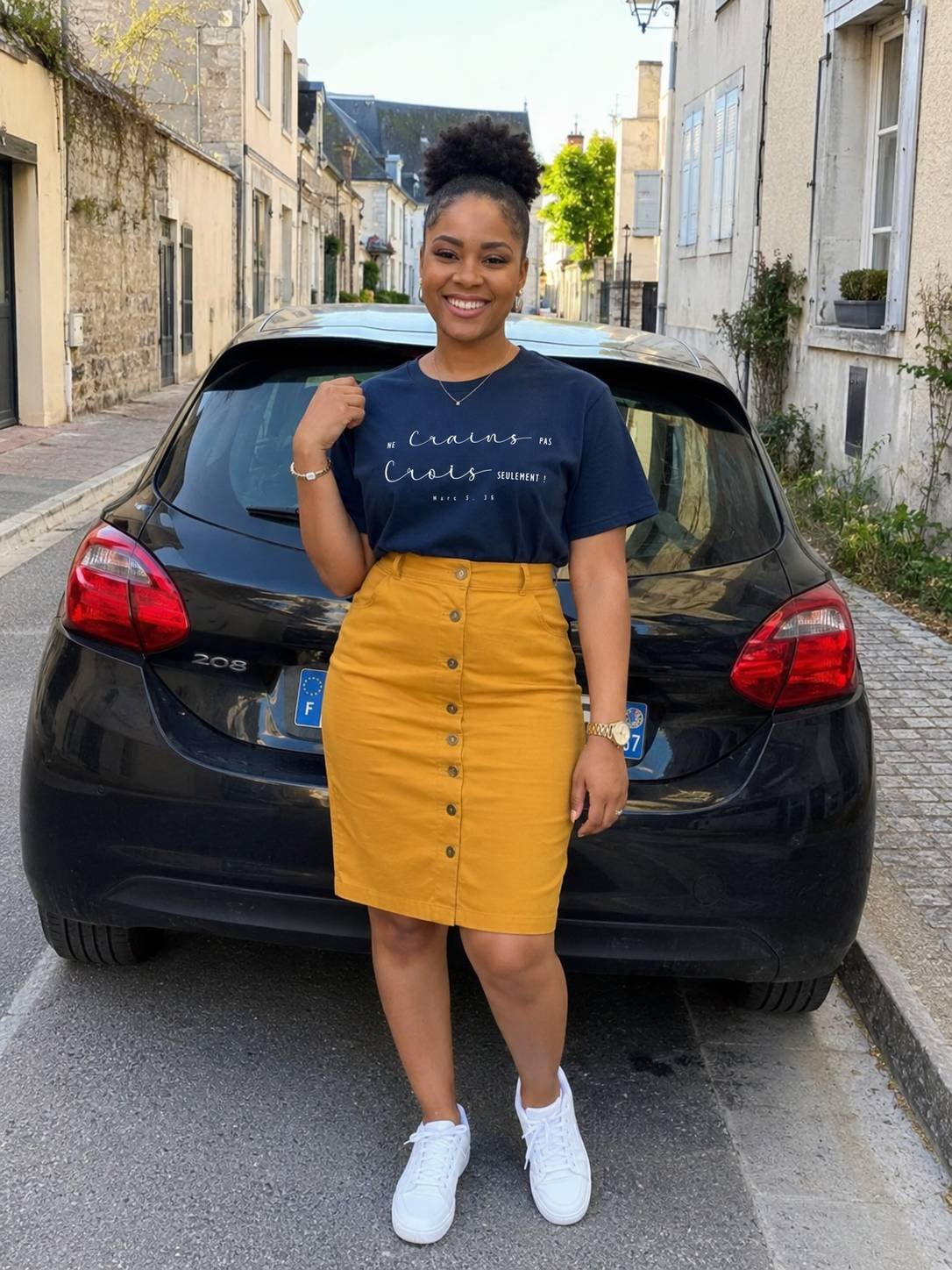 tshirtchretienfemmenecrainspasbleumarine Woman posing in a navy graphic T shirt mustard skirt and white sneakers on a cobblestone street beside a black hatchback smiling at the camera
