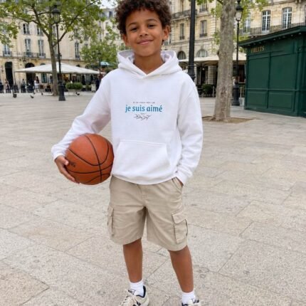 Boy standing in a sunny plaza, holding a basketball, wearing a white hoodie with 'je suis aimé' and beige cargo shorts.