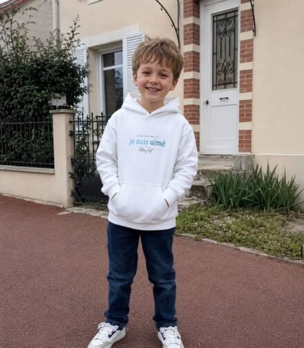 A smiling child stands on a red paved street in front of a beige house with brick accents, wearing a white hoodie and blue jeans with hands in the pockets.